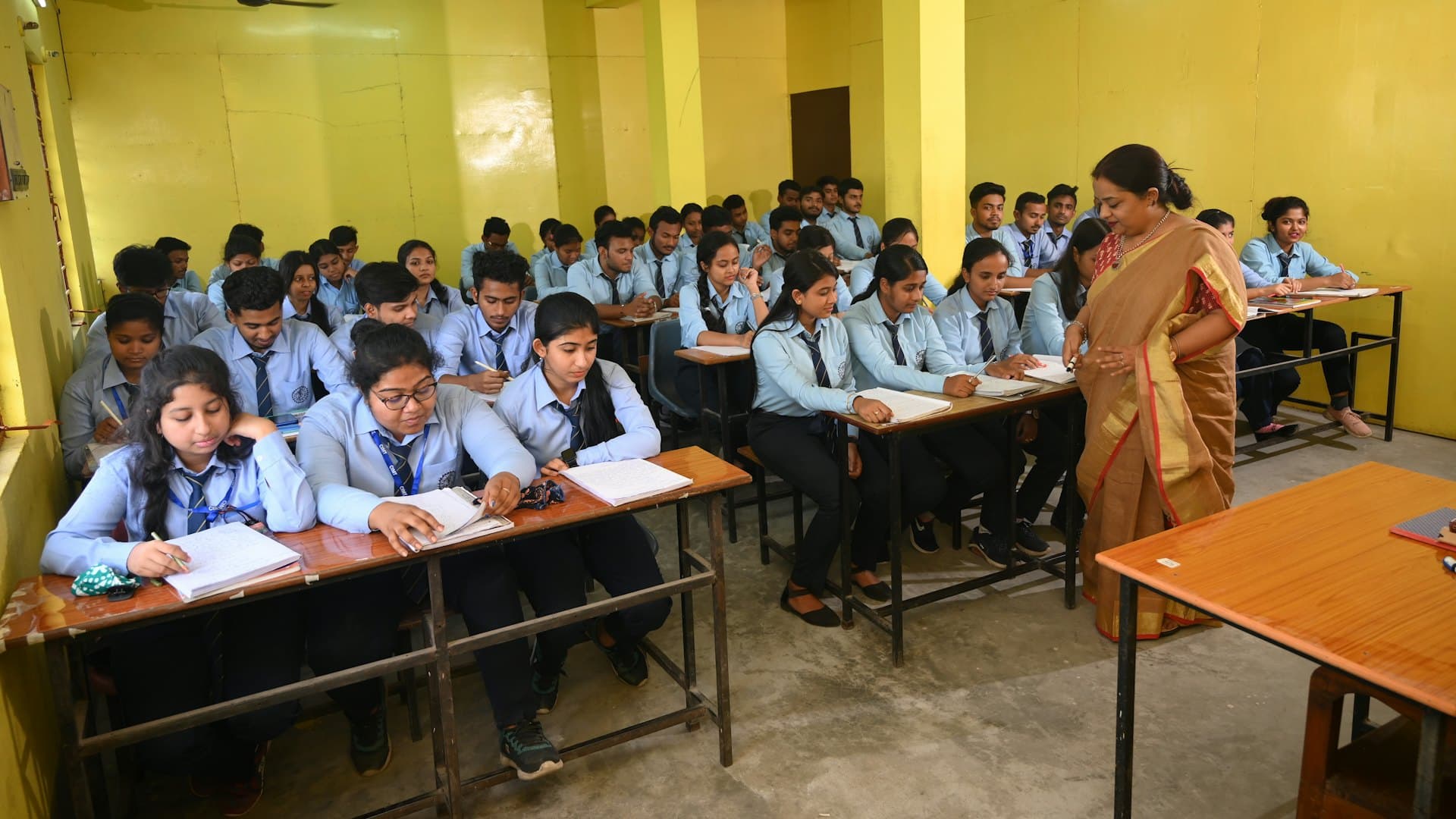 Indian classroom of uniformed students writing, teacher in saree assisting